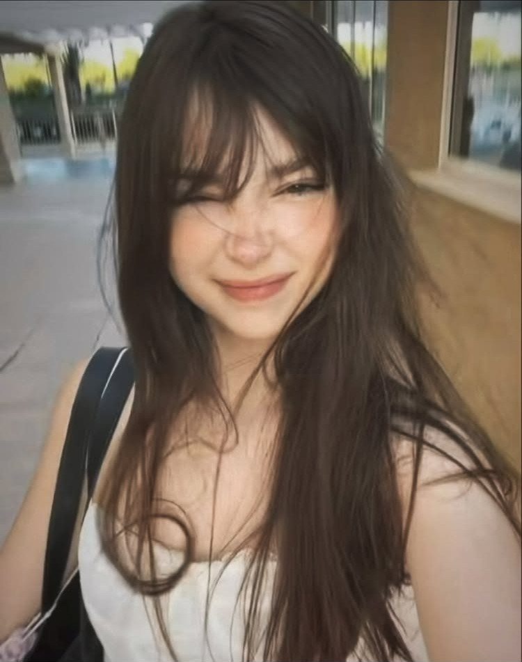 Young woman with long brown hair and wispy bangs smiling and winking.