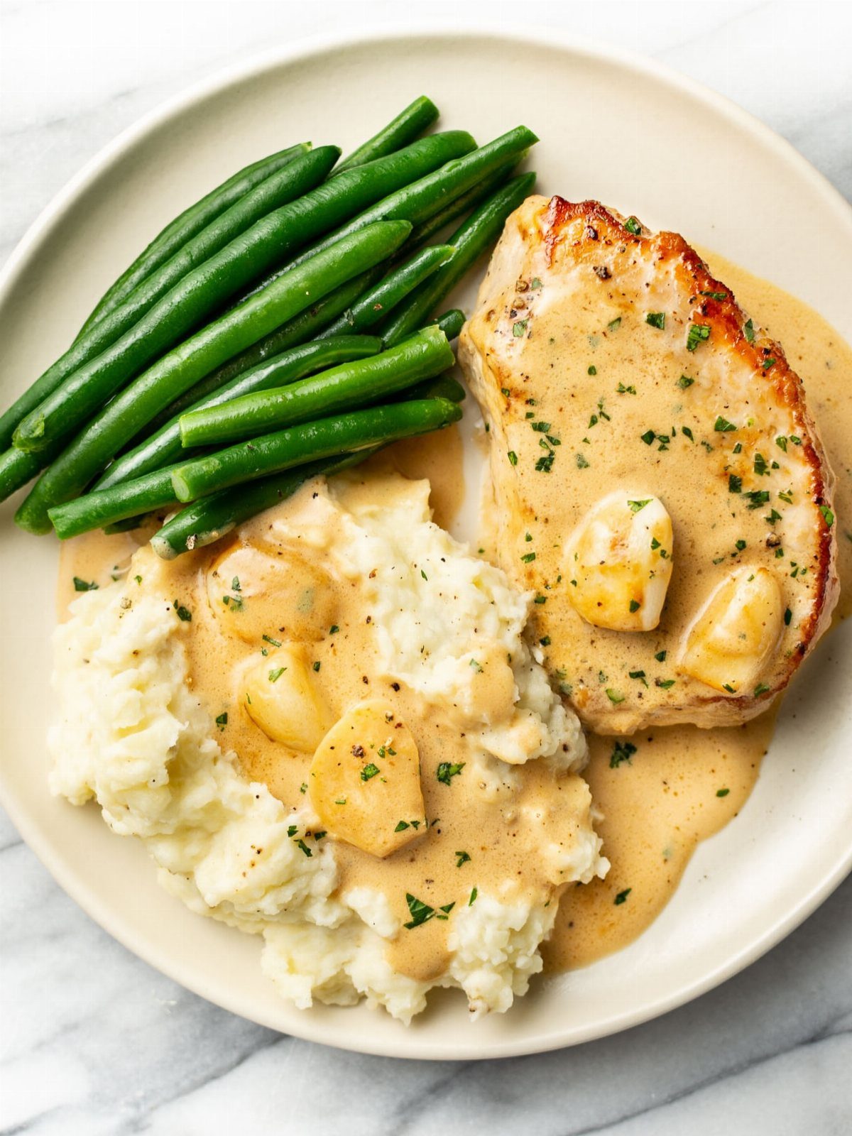 Cooked pork chop with creamy garlic sauce, mashed potatoes, and green beans on a plate.