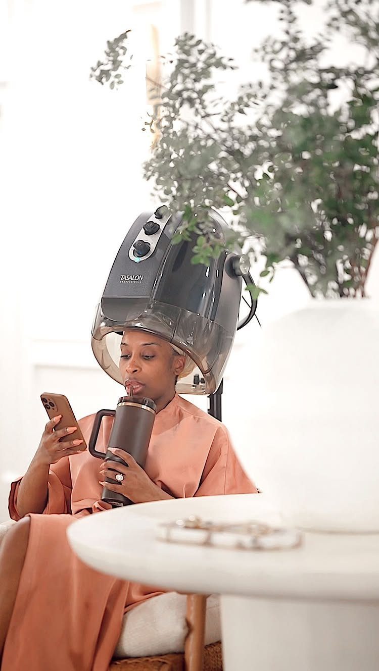 Woman under a hooded hair dryer holding a phone and a drinking tumbler.