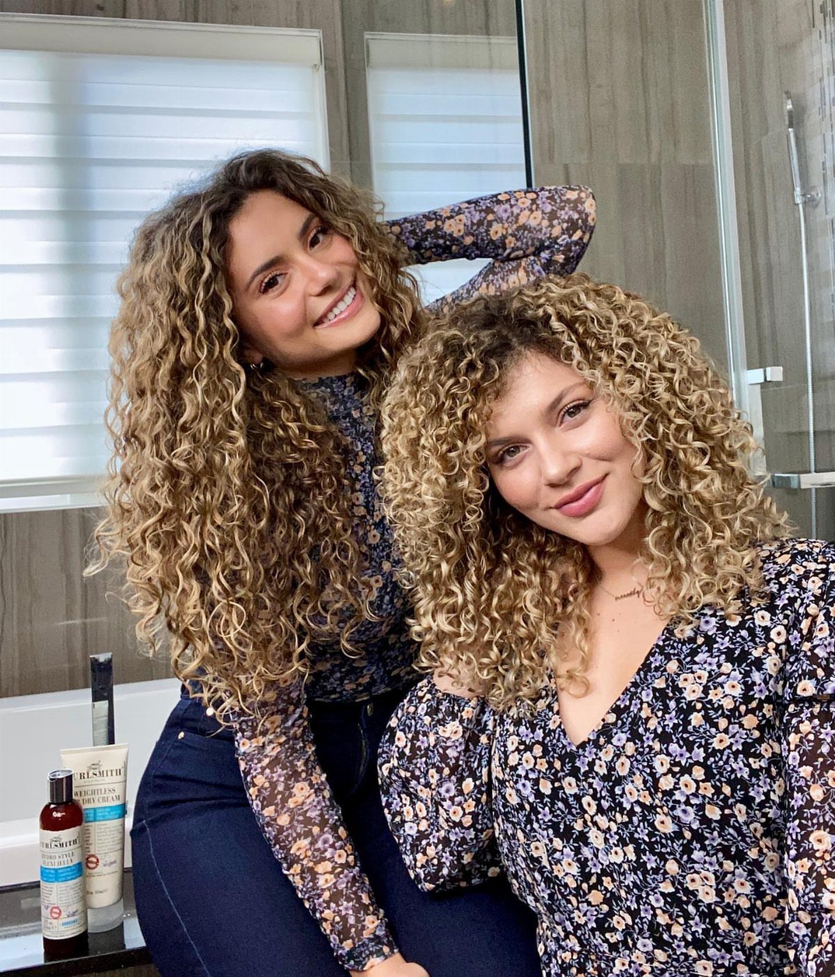Two women with voluminous curly hair posing in a bright bathroom setting.