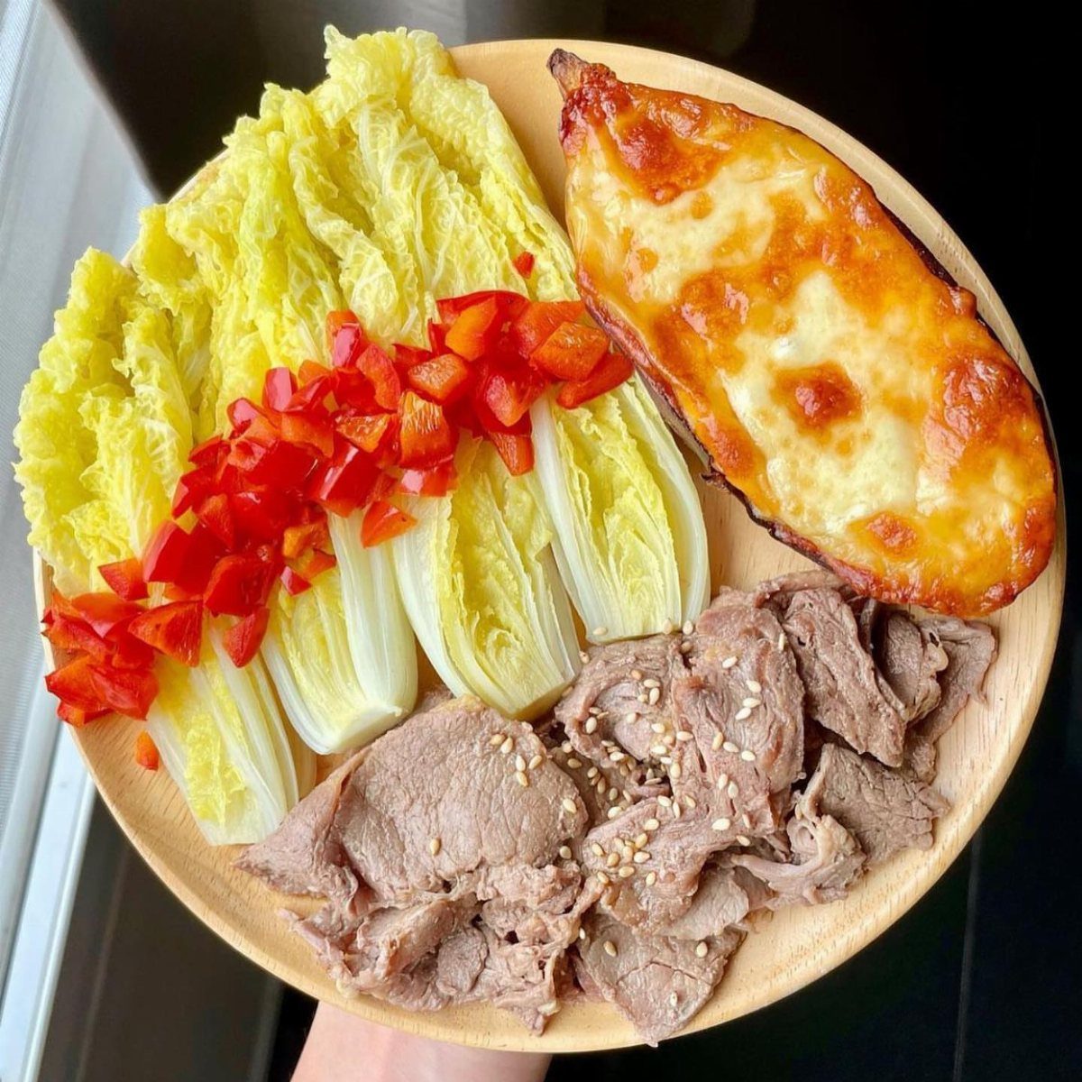 Wooden plate with sliced beef, steamed napa cabbage, red peppers, and cheesy baked vegetable.