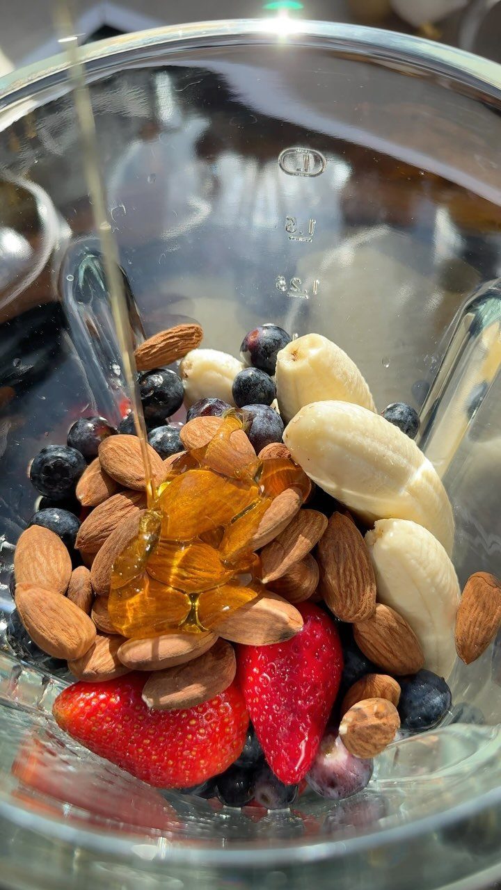 Blender jar filled with almonds, berries, and bananas with honey being poured over.