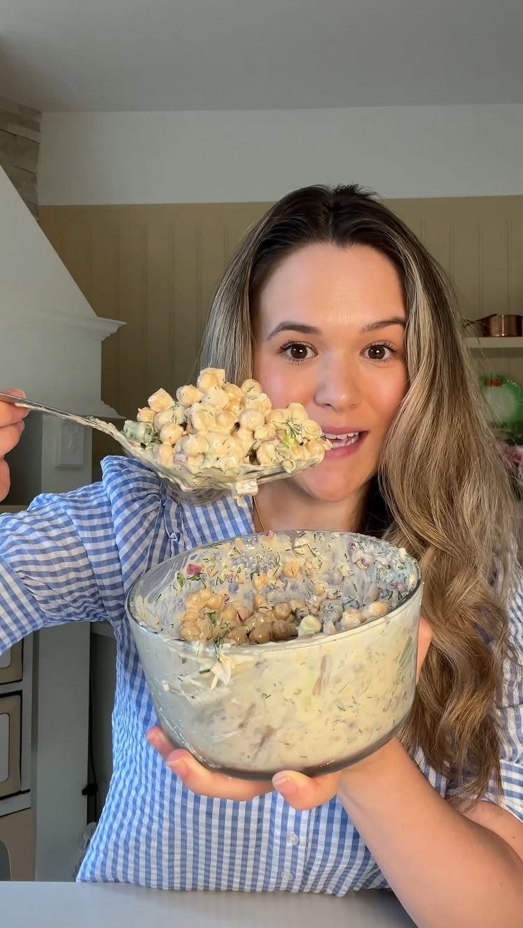 Woman holding a glass bowl and spoon of chickpea salad in a kitchen.
