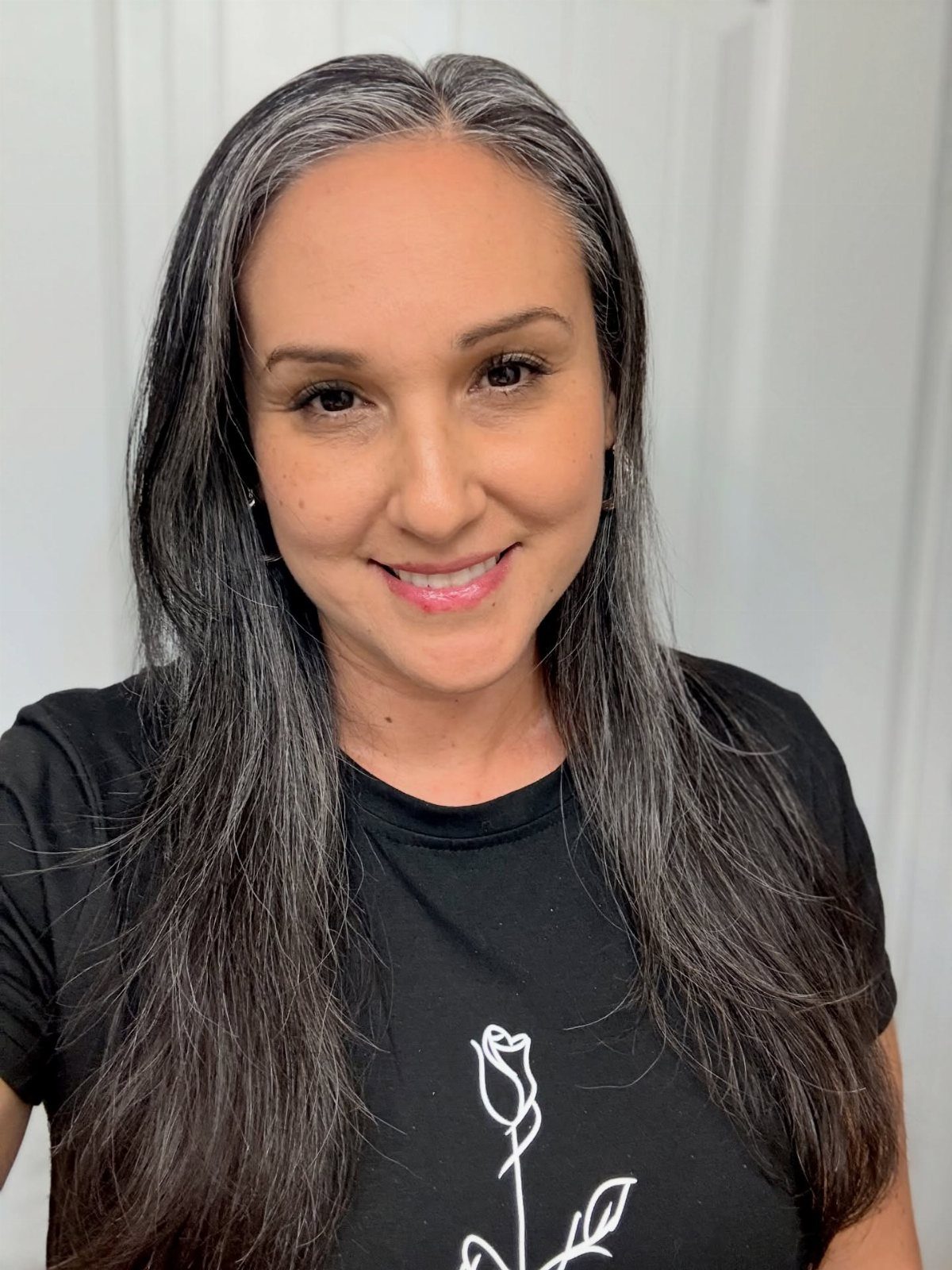 Woman with long dark hair and gray roots smiling against a white background