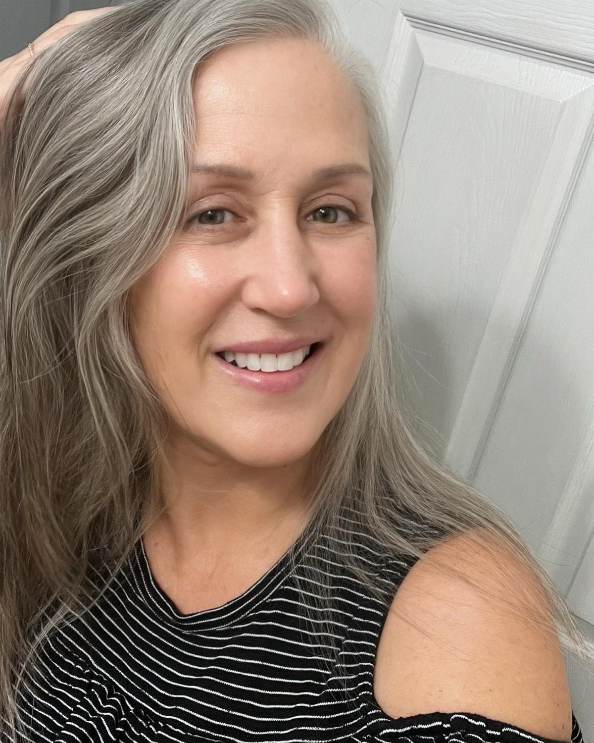 Woman with long natural gray hair smiling against a white paneled wall