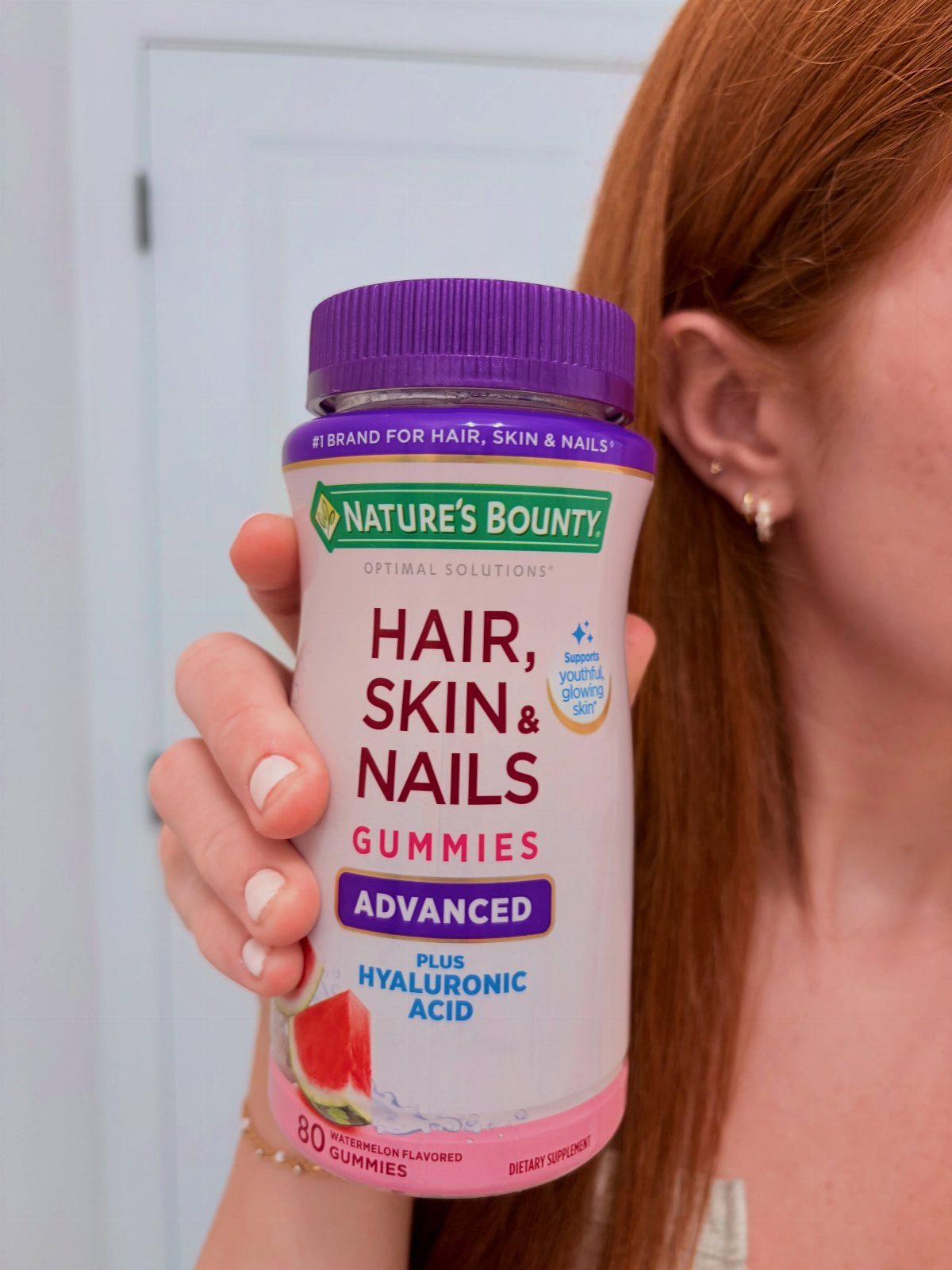 Nature's Bounty Hair, Skin & Nails Gummies bottle held by a person with red hair.