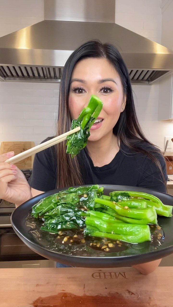 Woman eating sautéed green bok choy with chopsticks in a modern kitchen setting.