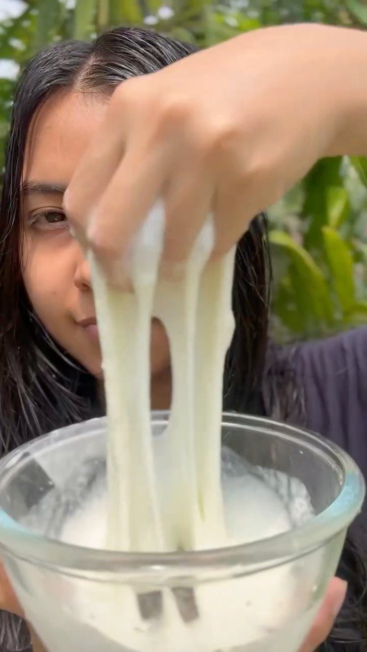 Woman pulling a stretchy white hair mask from a clear glass bowl outdoors.