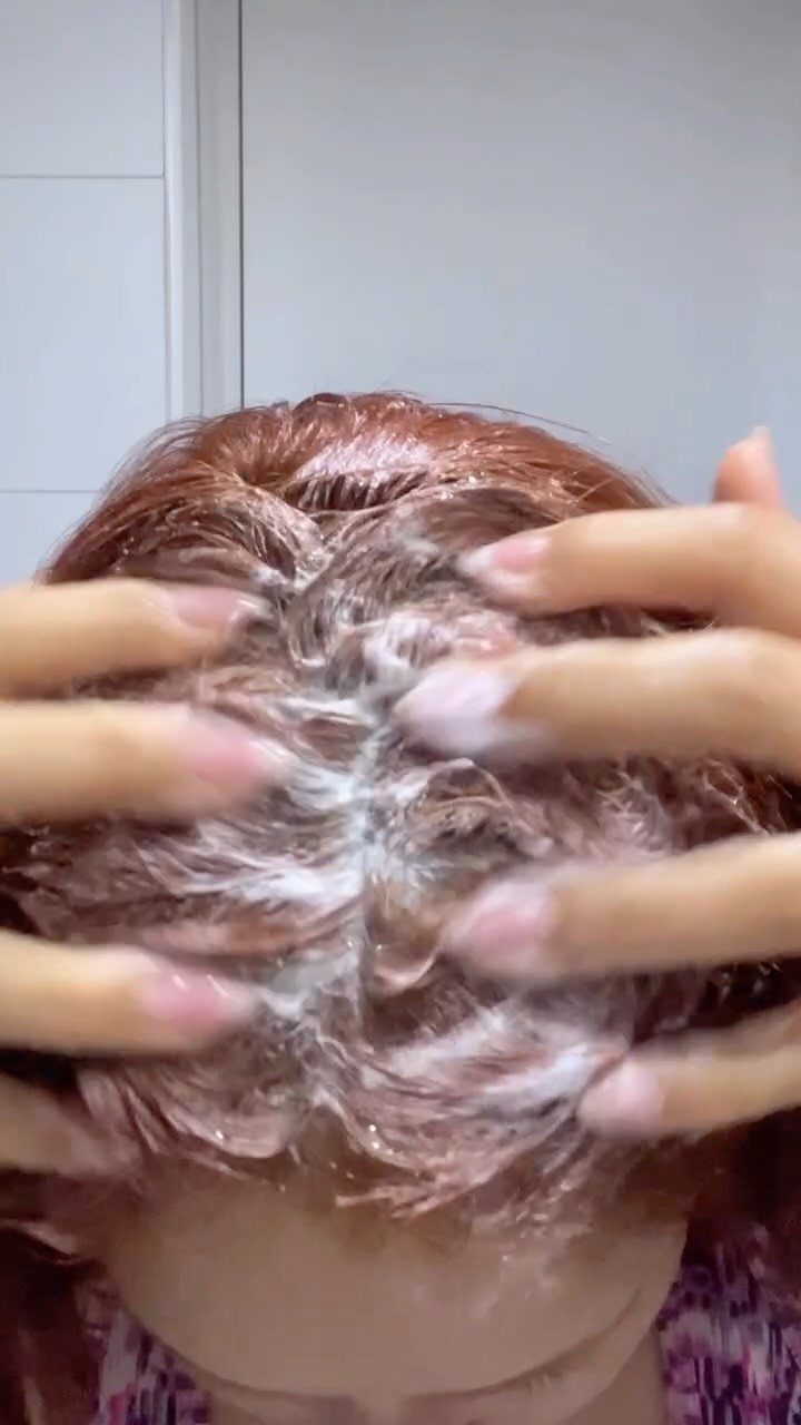 Person applying a white grainy scalp scrub to reddish-brown hair with fingers.