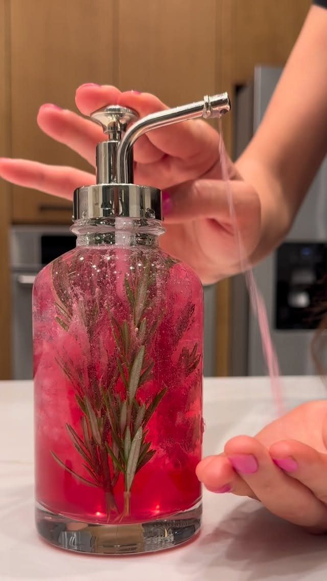 Pink grainy scrub with rosemary in a glass pump bottle being dispensed.