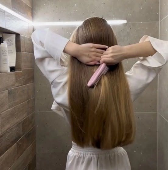 Woman with long thick light brown hair brushing it in a bathroom