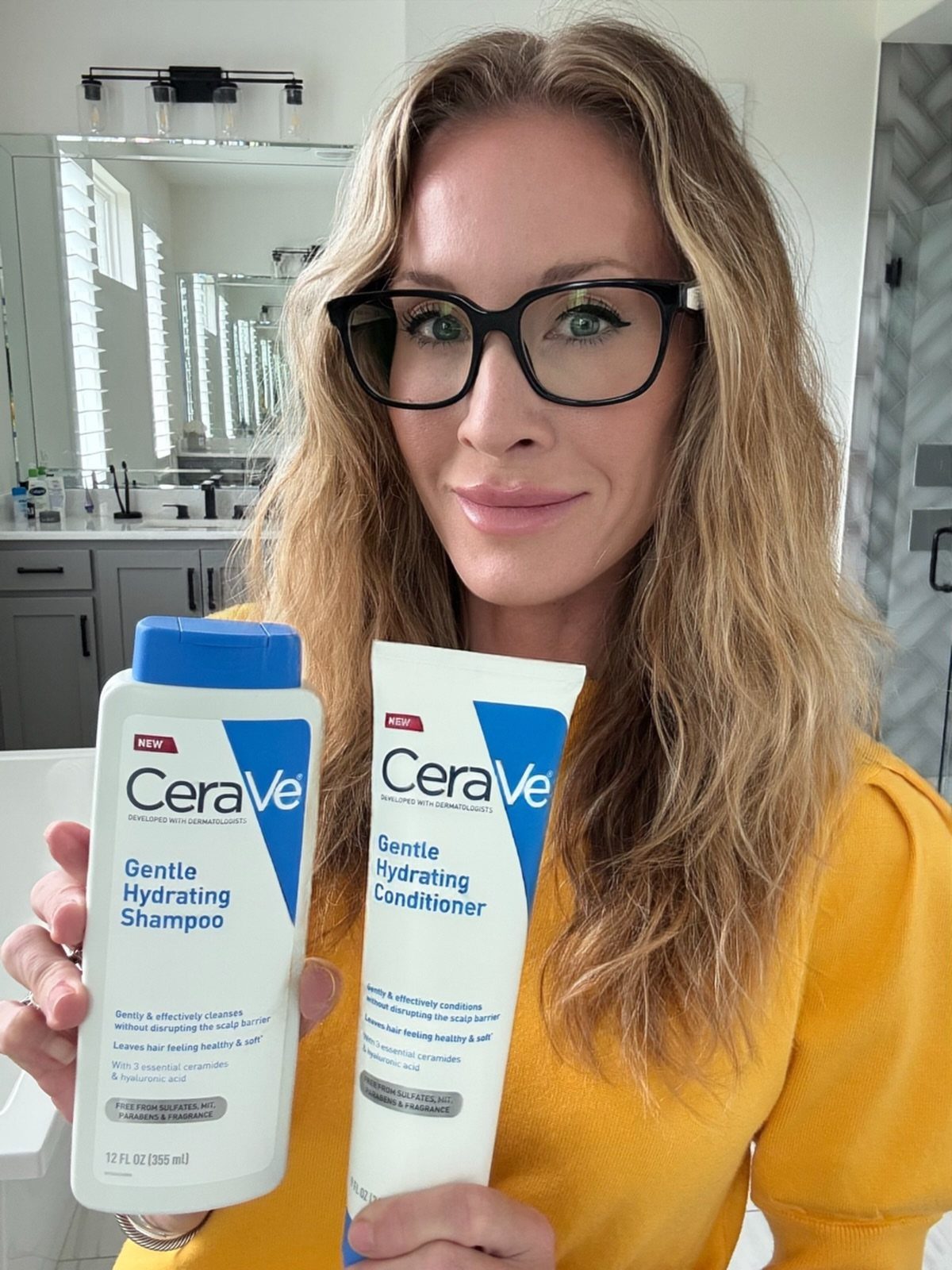 Woman holding CeraVe Gentle Hydrating Shampoo and Conditioner bottles in a bright bathroom.