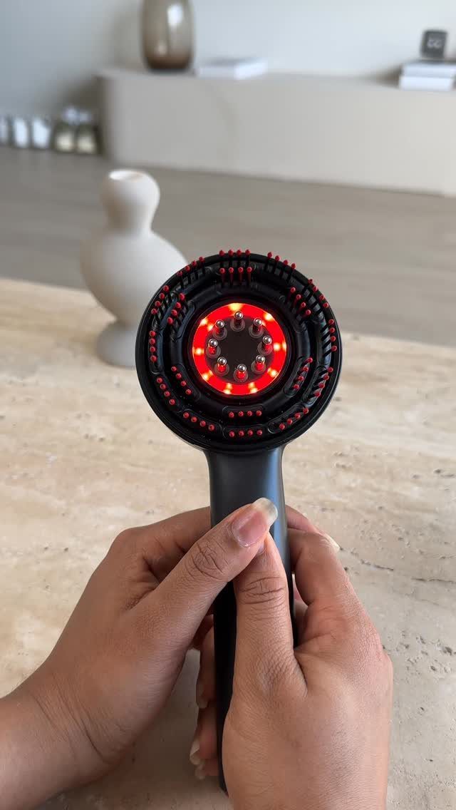 Black electric scalp massage brush with glowing red LED lights held by hands.