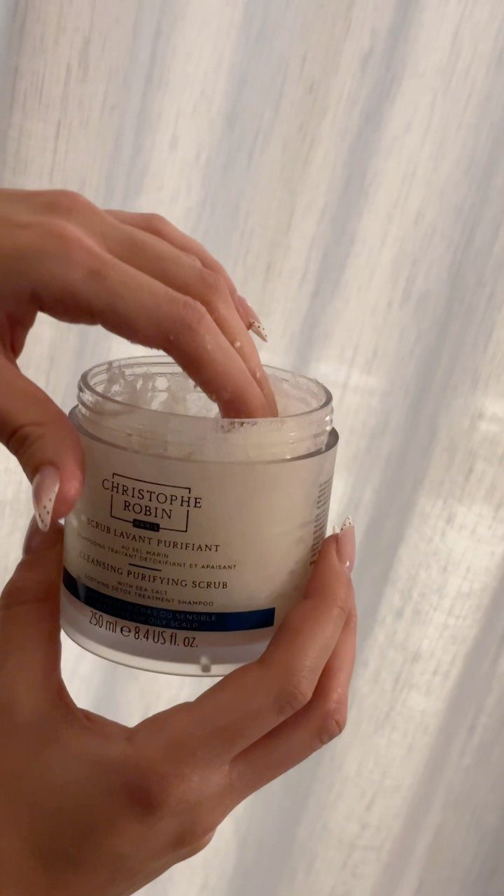 Hands holding a jar of Christophe Robin Cleansing Purifying Scrub with finger dipping inside.