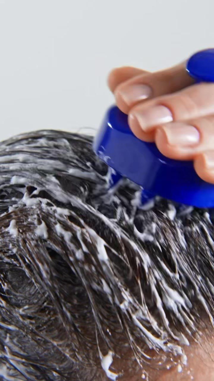 Blue scalp massaging brush scrubbing white soapy foam into dark wet hair
