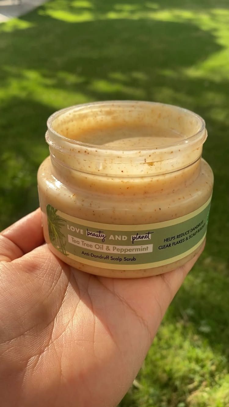Open jar of tea tree oil and peppermint scalp scrub held against green grass.