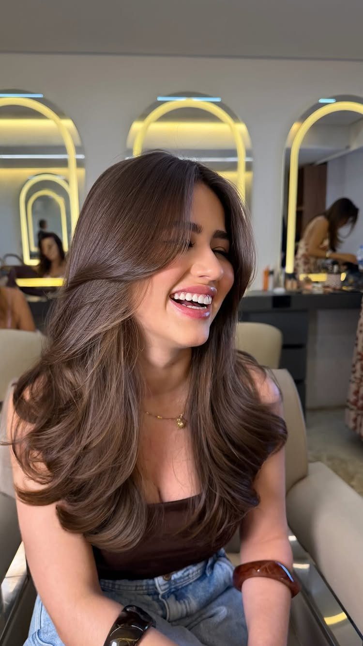 Woman with voluminous brown wavy hair and face-framing layers smiling in a salon.