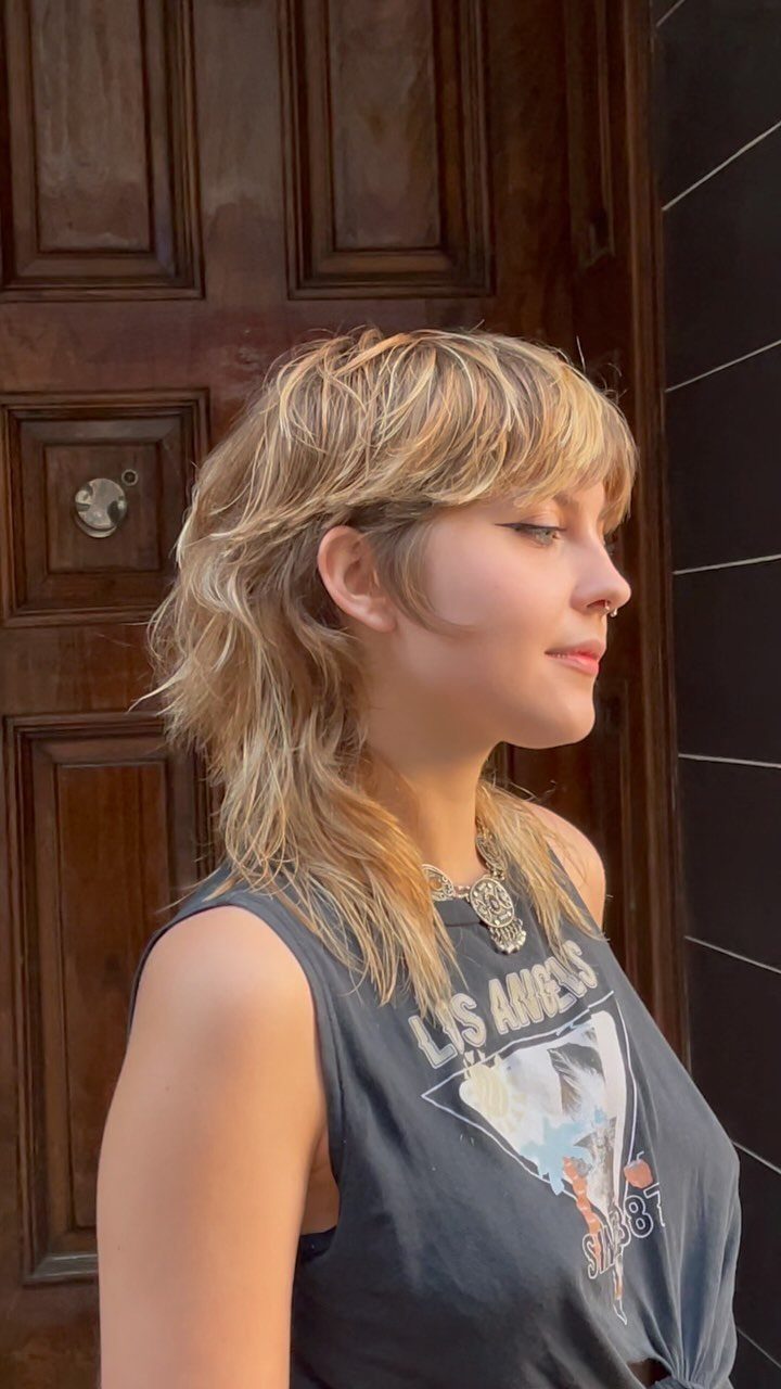 Young woman with a blonde soft wavy mullet haircut and bangs posing outdoors.