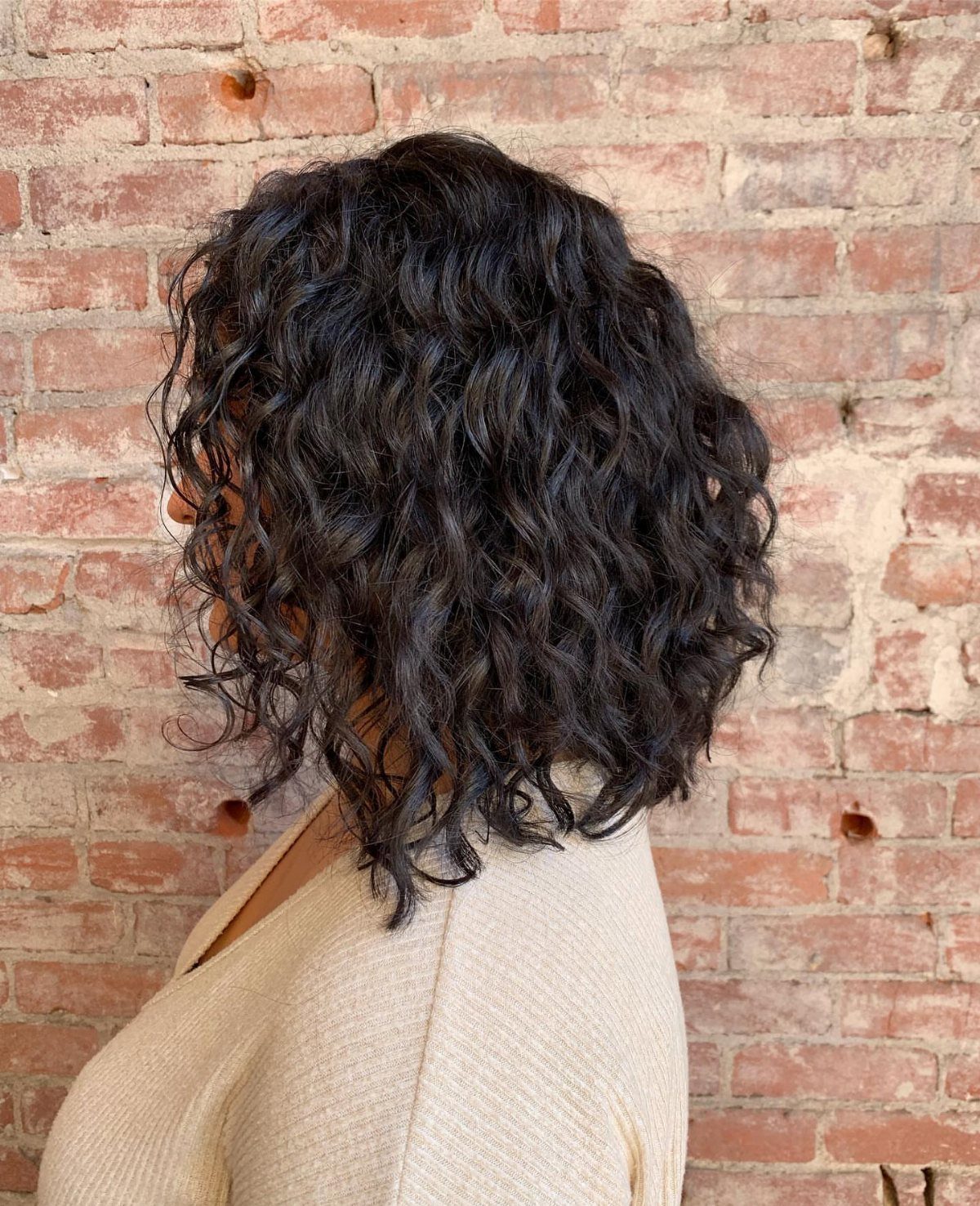 Curly dark brown shoulder-length hair against a rustic red brick wall background.